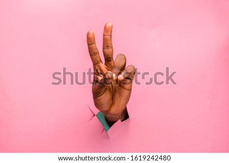 black person hand through a hole in a cardboard making v sign with two fingers, pink background