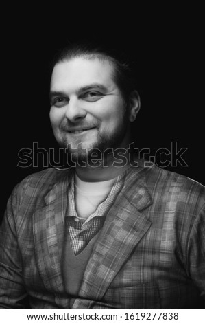 black and white low-key portrait of cheerful handsome bearded stylish elegant man in plaid blazer smiling and looking away on dark background