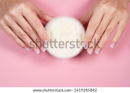 Closeup top view flatlay photography of beautiful manicured female hands and jar with moisturizing cream isolated on pink pastel background. Fingernails with classic french manicure. Horizontal photo.
