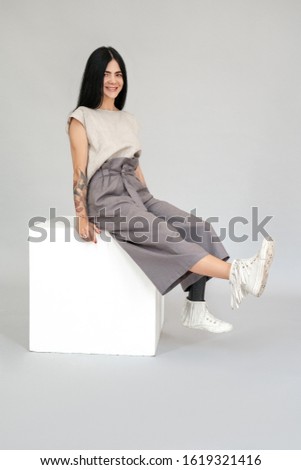 Diverse young woman with leg prosthesis sitting on white cube chair isolated on grey background shaking legs smiling joyful full body shot
