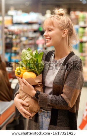Young woman at the supermarket standing holding paper bag with shoppings fresh organic vegetables looking aside laughing cheerful just walk out shopping technology Royalty-Free Stock Photo #1619321425