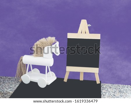 Mock up in black, white and purple colors, with black empty surface for text on the small easel chalkboard and decorated with white little wooden horse toy by side on the foggy purple background