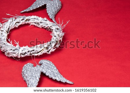 A door wreath of branches decorated with artificial snow. Nearby are angel wings of silver color. Everything stands on a red background with a place for text. Top views