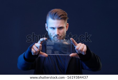 Young man with beard and trendy hairstyle uses futuristic smartphone with transparent display. Future technology, innovative ideas concept. Blue neon light. Free space for text. Dark background.