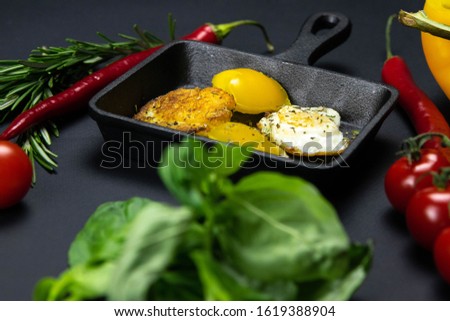 A small black frying pan with quail eggs in the shape of a heart on the blacl background, next to it are caenese pepper, bell pepper tomatoes, rosemary and basil. Top views
