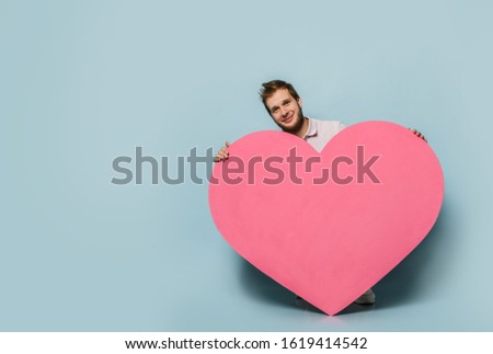 Young man standing with pink heart shape toy in hands on pastel blue wall background. Love gift for valentines day with text space