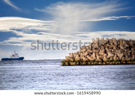 The seawall around the sea port facilities is made of concrete tetrapods (traveling-wave protection), rubble-mound breakwater. Duty marine tug. Unusual clouds over autumn sea and fedeeng marine ducks Royalty-Free Stock Photo #1619458990