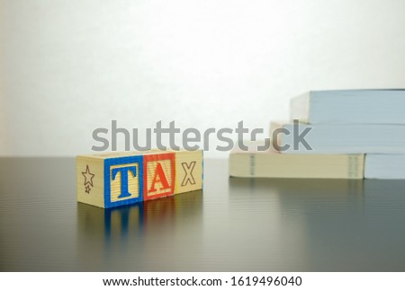 Conceptual image of annual tax return. Wooden block letter forming TAX besides stacked books. White background and dark reflective surface. Focus at block letter T.