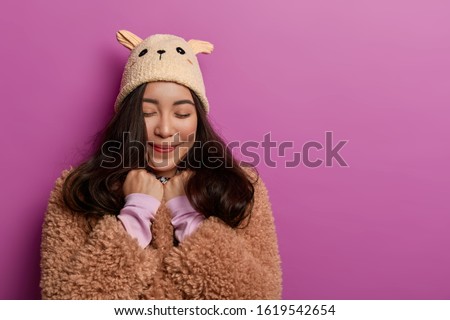 Studio shot of pleasant looking Asian woman pleased hear heartwarming compliment, keeps eyes closed, holds hands clenched in fists near face wears winter outerwear, stands indoor over vivid background