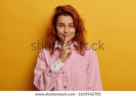 Playful redhead pretty woman with satisfied secret look, makes hush gesture, gossips with friend, dressed in rosy denim jacket, hints on secret, isolated over yellow background. Dont tell anyone