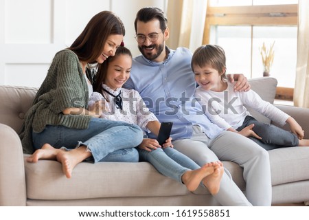 Overjoyed young parents sit rest on comfortable sofa in living room with small kids watch funny video on smartphone together, happy family with little children relax on couch at home use modern cell