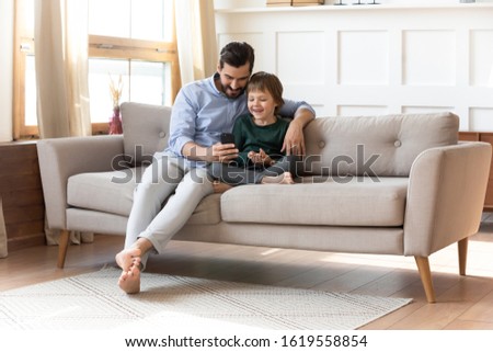 Caring young dad sit relax on couch in living room with little preschooler son watch funny video on cellphone together, smiling father rest on sofa have fun play use smartphone with small boy child