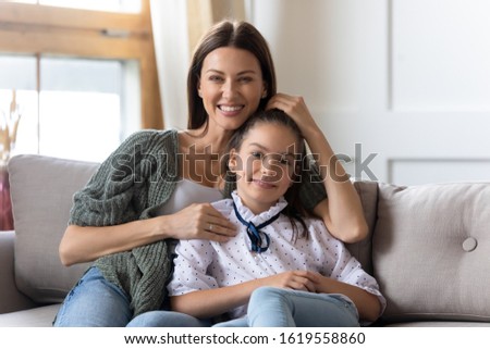 Portrait of happy young mother sit on couch in living room hug embrace cute little daughter, smiling mom or nanny cuddle small girl child look at camera posing enjoy family weekend time together