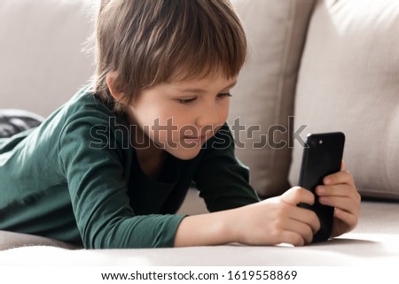 Cute smart little boy lying on couch in living room playing game on modern cellphone gadget, small kid have fun watch video or cartoon on smartphone at home, children and technology concept