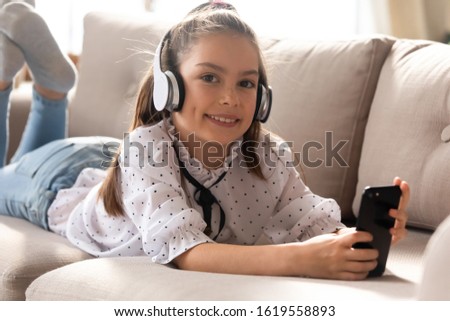 Portrait of smiling cute little girl lying on couch wear modern headphones using smartphone at home, happy small kid in earphones watch cartoons play browse Internet on modern cellphone gadget