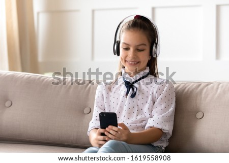 Smiling little girl sit on couch in living room wear headphones watch video on cellphone, happy small kid in earphones relax on sofa have fun using smartphone at home, children and technology concept