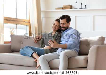 Happy young couple sit relax on couch in living room watch movie on tablet together, loving millennial husband and wife rest on cozy sofa at home using modern pad gadget, family weekend concept Royalty-Free Stock Photo #1619558911