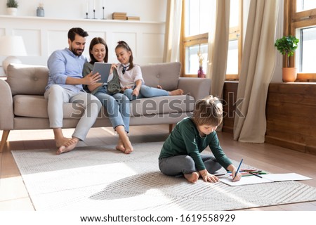 Small preschooler boy kid sit on floor drawing painting picture in album, young Caucasian parents and little girl daughter relax on couch using tablet together, happy family weekend at home