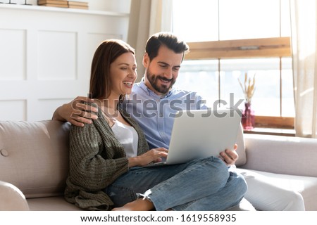 Happy millennial couple sit relax on couch in living room watching video on laptop together, smiling young husband and wife rest on sofa at home browsing Internet using modern computer device Royalty-Free Stock Photo #1619558935
