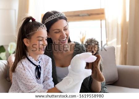 Excited young mom or nanny sit on couch play with cute girl child with hand toys puppet dolls, overjoyed mother relax have fun with small daughter engaged in funny activity in living room at home