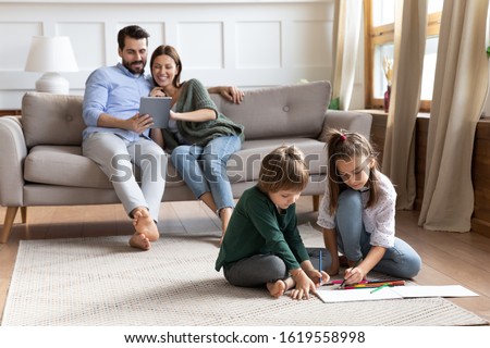 Cute little brother and sister sit on floor drawing in album, young parents relax on comfortable couch in living room using tablet, young Caucasian family rest together enjoy leisure weekend at home