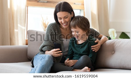 Caring young mum sit on couch in living room with small son watch funny cartoon on smartphone, happy mother or nanny relax on sofa at home with little boy child use cell browse Internet together