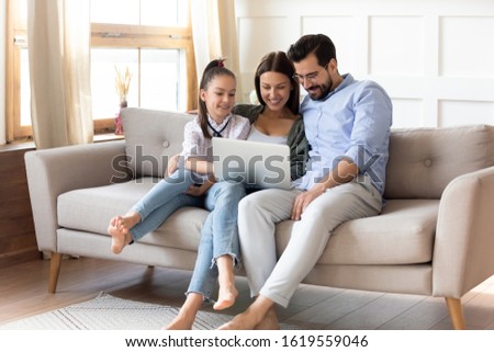 Smiling young parents sit relax on comfortable sofa in living room with little daughter watch video on laptop together, happy family with small girl child rest enjoy weekend at home using computer