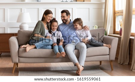 Happy young Caucasian family with little kids sit relax on comfortable sofa in living room using smartphone together, smiling parents rest with small children on couch at home browsing cellphone