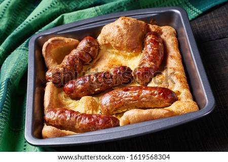Classic british dish toad in the hole of yorkshire pudding with roasted sausages served on a square cake pan on a dark rustic wooden background, horizontal view, close-up
