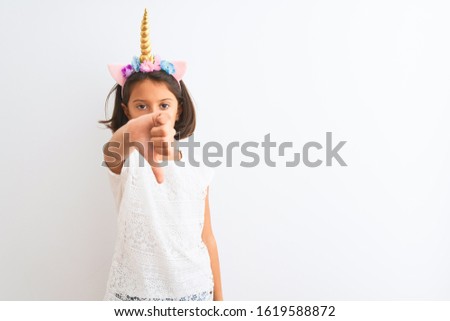 Beautiful child girl wearing unicorn diadem standing over isolated white background looking unhappy and angry showing rejection and negative with thumbs down gesture. Bad expression.