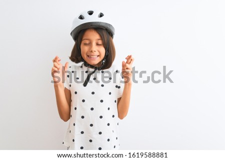 Beautiful child girl wearing security bike helmet standing over isolated white background gesturing finger crossed smiling with hope and eyes closed. Luck and superstitious concept.