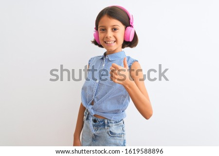 Beautiful child girl listening to music using headphones over isolated white background doing happy thumbs up gesture with hand. Approving expression looking at the camera with showing success.