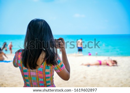 The back of a dark skinned girl who wearing a bright-colored shirt, She is using a smartphone to take pictures of the beach and sea scenery with tourists who are playing in the water And sunbathe.