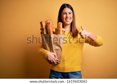 Young beautiful woman holding a bag of fresh healthy bread over yellow background doing happy thumbs up gesture with hand. Approving expression looking at the camera showing success.