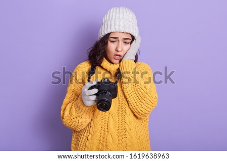 Portrait of shocked attractive emotional brunette holding photocamera in one hand, looking through photos, unsatisfied with results, being disappointed with quality. People and emotions concept.