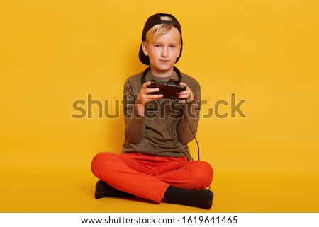 Little blond boy wearing casaul clothing, holding in hand modern device, playing game on mobile phone isolated over yellow background, looking directly at camera. Kids, childhood, lifestyle concept.
