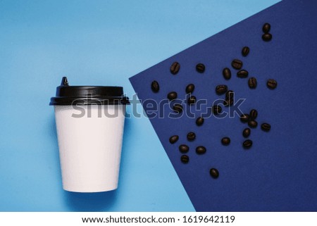 White cardboard glass with a plastic black lid on a blue and cyan background with coffee beans. Glass with take-away coffee. The trend color is classic blue. Copyspace, flat lay.