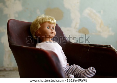 Old plastic doll with blonde hair sitting in old-fashioned crimson leather chair.