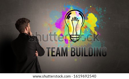 businessman drawing colorful light bulb with TEAM BUILDING inscription on textured concrete wall, new business idea concept