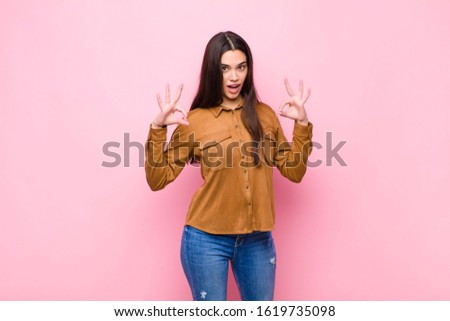 young pretty woman feeling shocked, amazed and surprised, showing approval making okay sign with both hands against pink wall