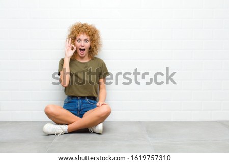 young afro woman feeling successful and satisfied, smiling with mouth wide open, making okay sign with hand sitting on a floor