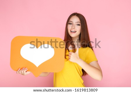 Young woman holding paper card with heart and showing thumb up on pink background
