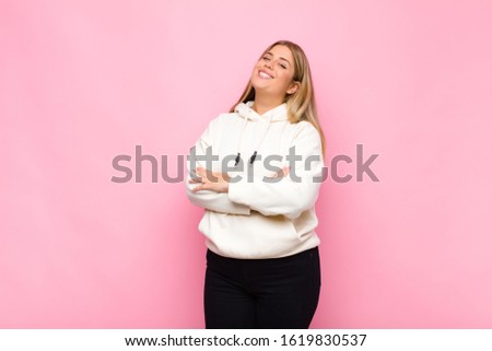 young blonde woman laughing happily with arms crossed, with a relaxed, positive and satisfied pose against flat wall