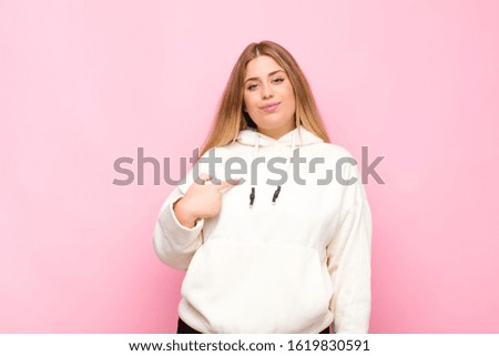 young blonde woman looking proud, confident and happy, smiling and pointing to self or making number one sign against flat wall