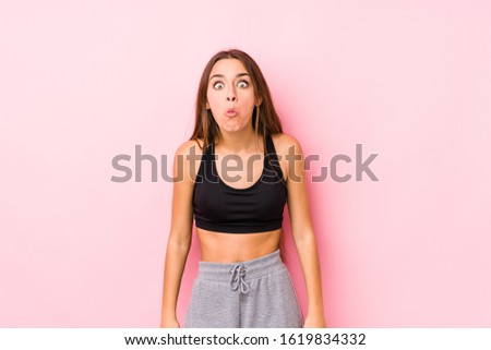 Young caucasian fitness woman posing in a pink background shrugs shoulders and open eyes confused.