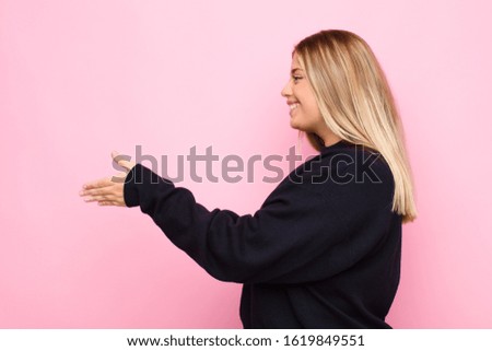 young blonde woman smiling, greeting you and offering a hand shake to close a successful deal, cooperation concept against flat wall