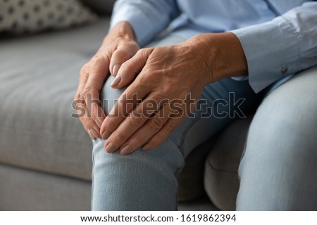 Close up older woman massaging touching knee with hands, feeling pain, mature female suffering from ache in leg, sitting on couch at home, feeling unwell, health problem concept, osteoarthritis Royalty-Free Stock Photo #1619862394