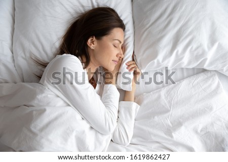 Serene beautiful woman sleeping with hand under cheek on soft pillow, young female wearing white pajamas resting under warm blanket in comfortable bed in bedroom or in hotel, enjoying sweet dreams Royalty-Free Stock Photo #1619862427