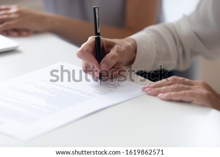 Close up view female hand holding pen signing legal paper seated at desk indoors, client receives services affirming document, hiring and human resources concept, put signature on employment contract