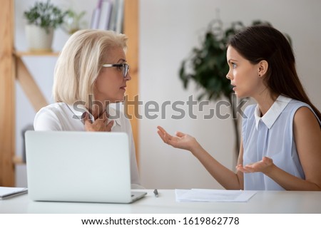 Middle-aged and millennial female colleagues having different opinions on joint project arguing seated at workplace, negative attitude, inequality and discrimination, poor work environment concept Royalty-Free Stock Photo #1619862778